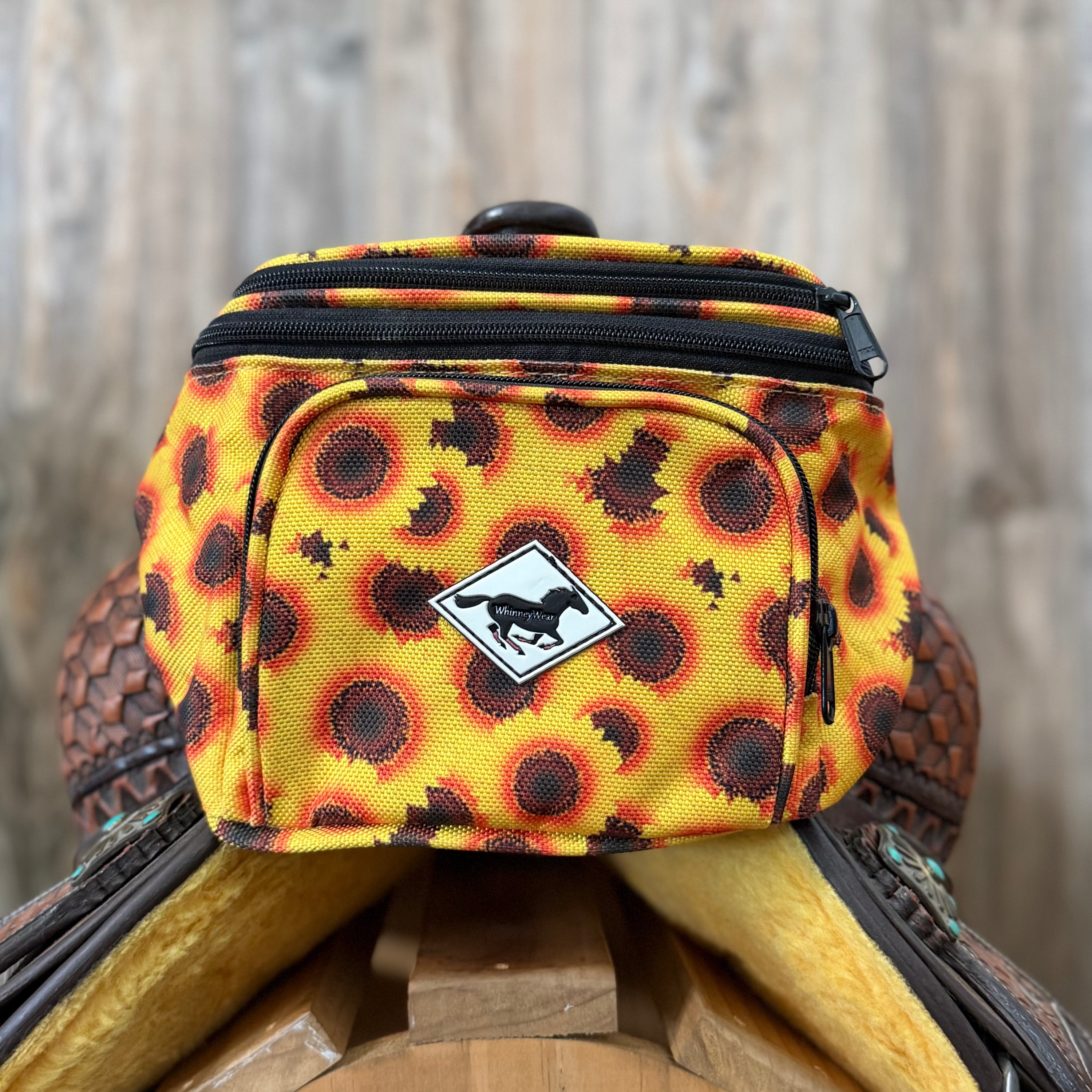 A small yellow and black sunflower-patterned fanny pack with a white logo patch, resting on a wooden barrel against a rustic wood wall background.